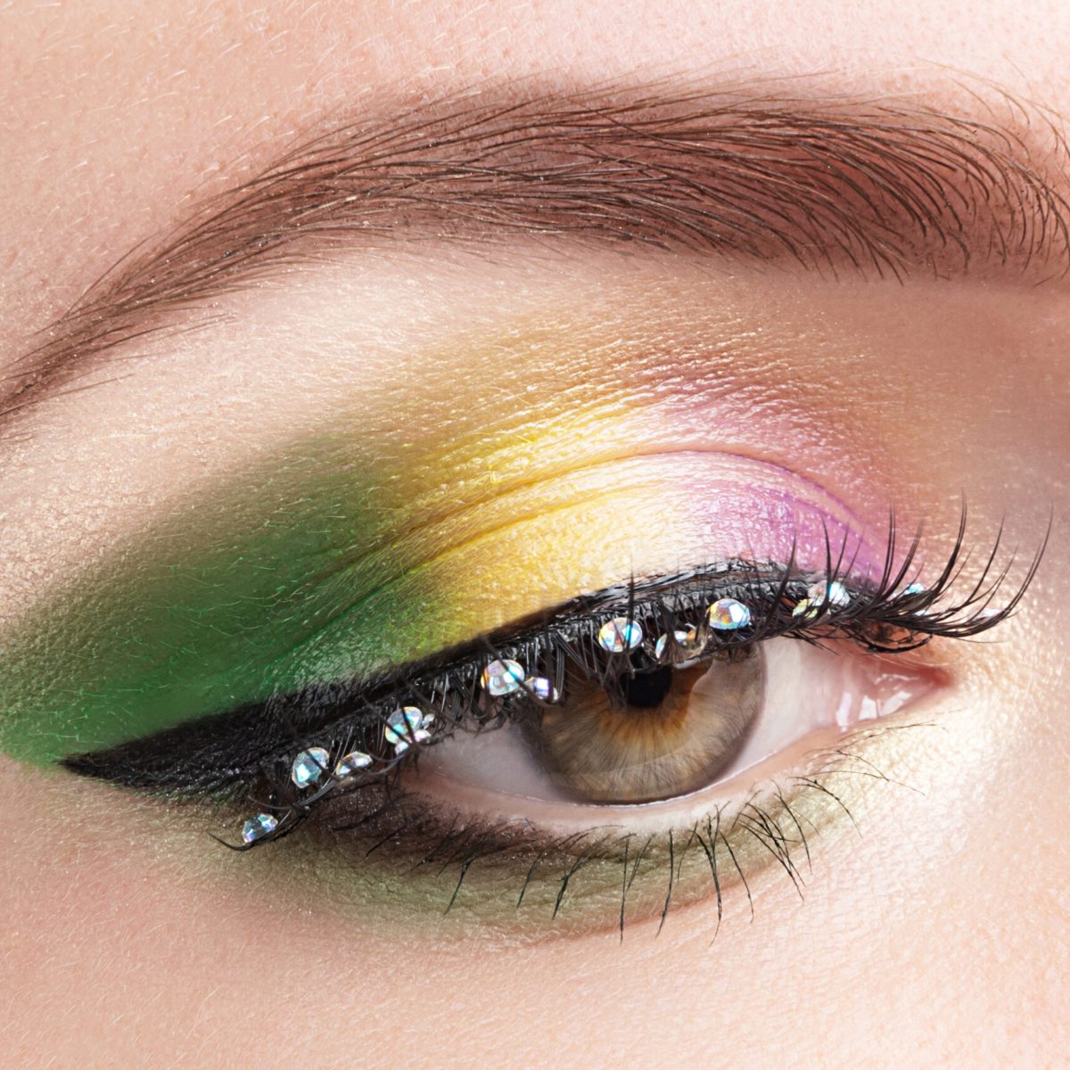 Gold, purple and green Mardi gras makeup. Woman closed eye with bright eyeshadows and rhinestones on false eyelashes closeup Gold, purple and green Mardi gras makeup. Woman closed eye with bright eyeshadows and rhinestones on false eyelashes closeup.